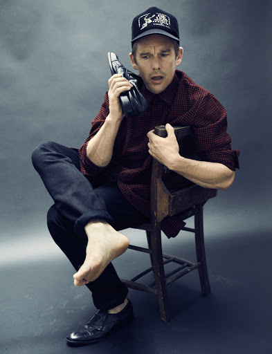 Ethan Hawke holding a leather shoe to his ear like a phone while seated backwards on a wooden chair against a grey background.