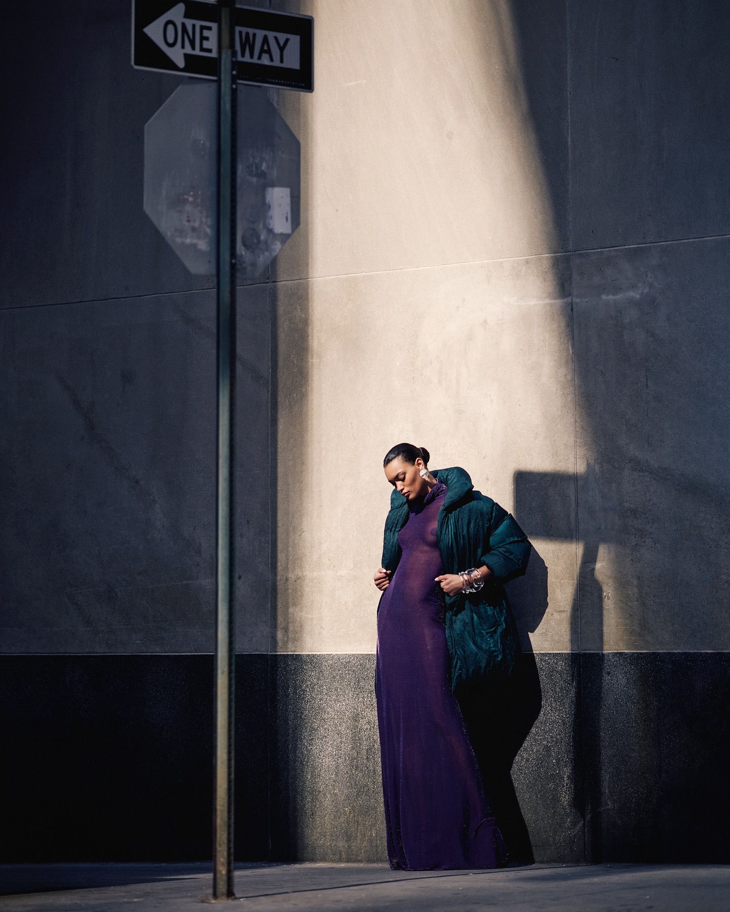 Woman in a long purple dress and green puffer jacket standing next to a One Way sign on a city street on a sunny day.