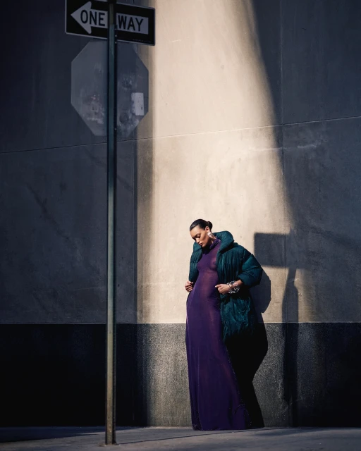Woman in a long purple dress and green puffer jacket standing next to a One Way sign on a city street on a sunny day.