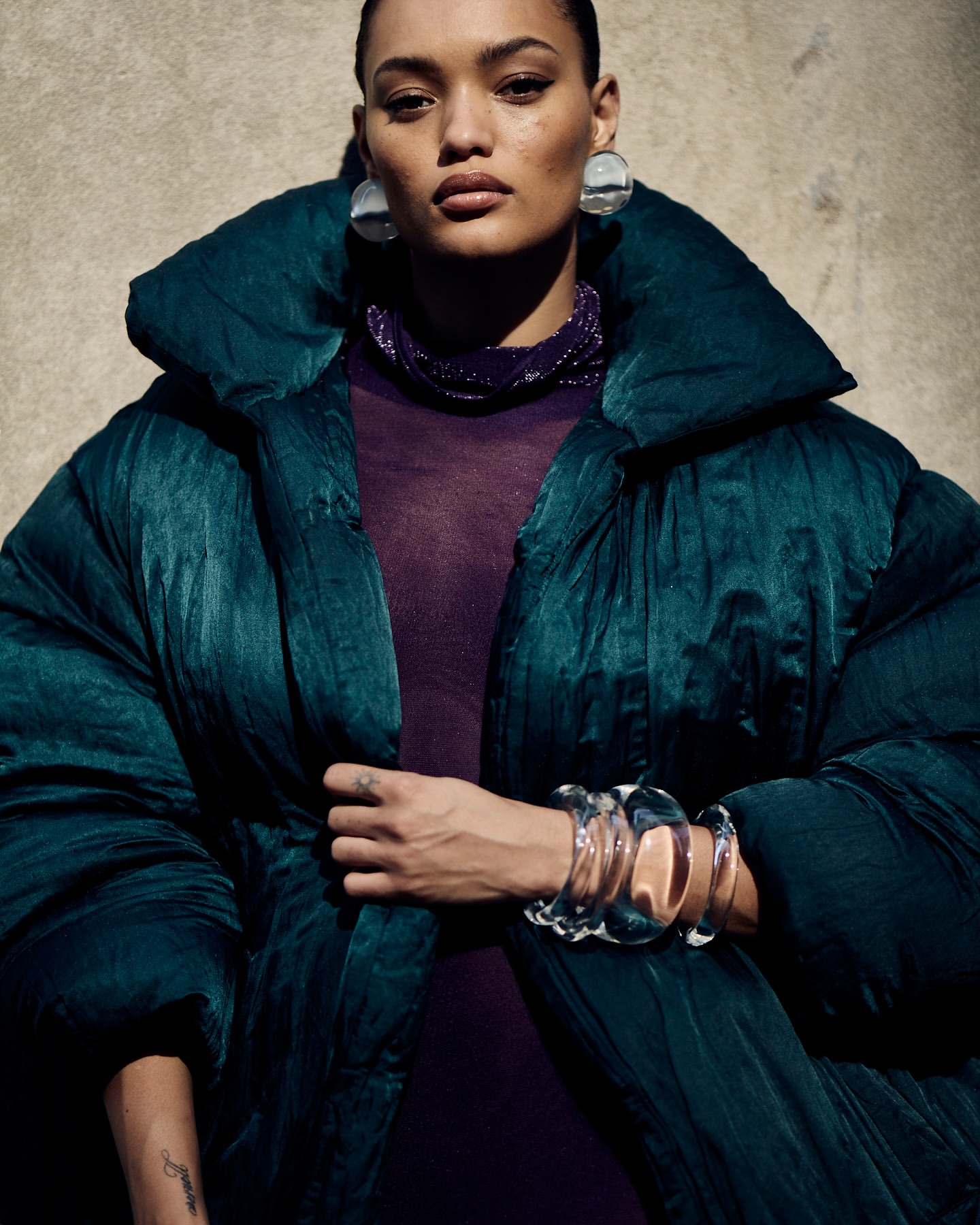 Elegant adult woman wearing a dark green puffer coat and multiple transparent bangle bracelets, posing in front of a textured wall.