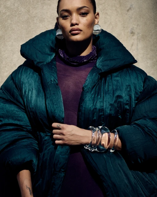 Elegant adult woman wearing a dark green puffer coat and multiple transparent bangle bracelets, posing in front of a textured wall.