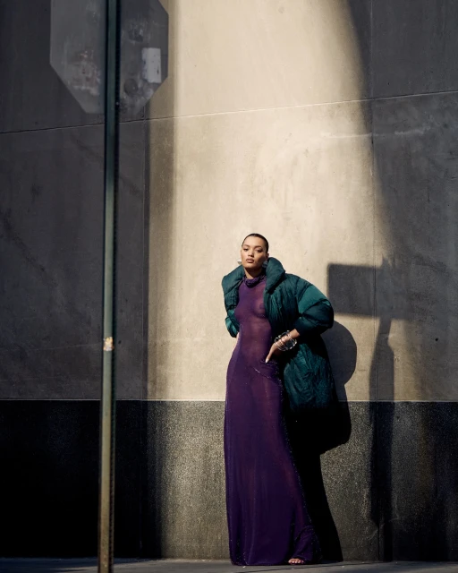 A woman poses outdoors in a sheer purple dress and a green puffer jacket, standing against a textured concrete wall on a sunny day.