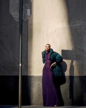 A woman poses outdoors in a sheer purple dress and a green puffer jacket, standing against a textured concrete wall on a sunny day.