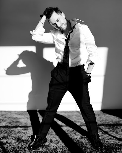 Black-and-white portrait of a man in dress shirt and dress pants, with a hand on his head, shadow visible on a neutral wall.