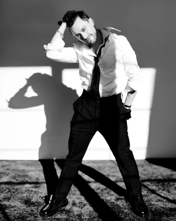 Black-and-white portrait of a man in dress shirt and dress pants, with a hand on his head, shadow visible on a neutral wall.