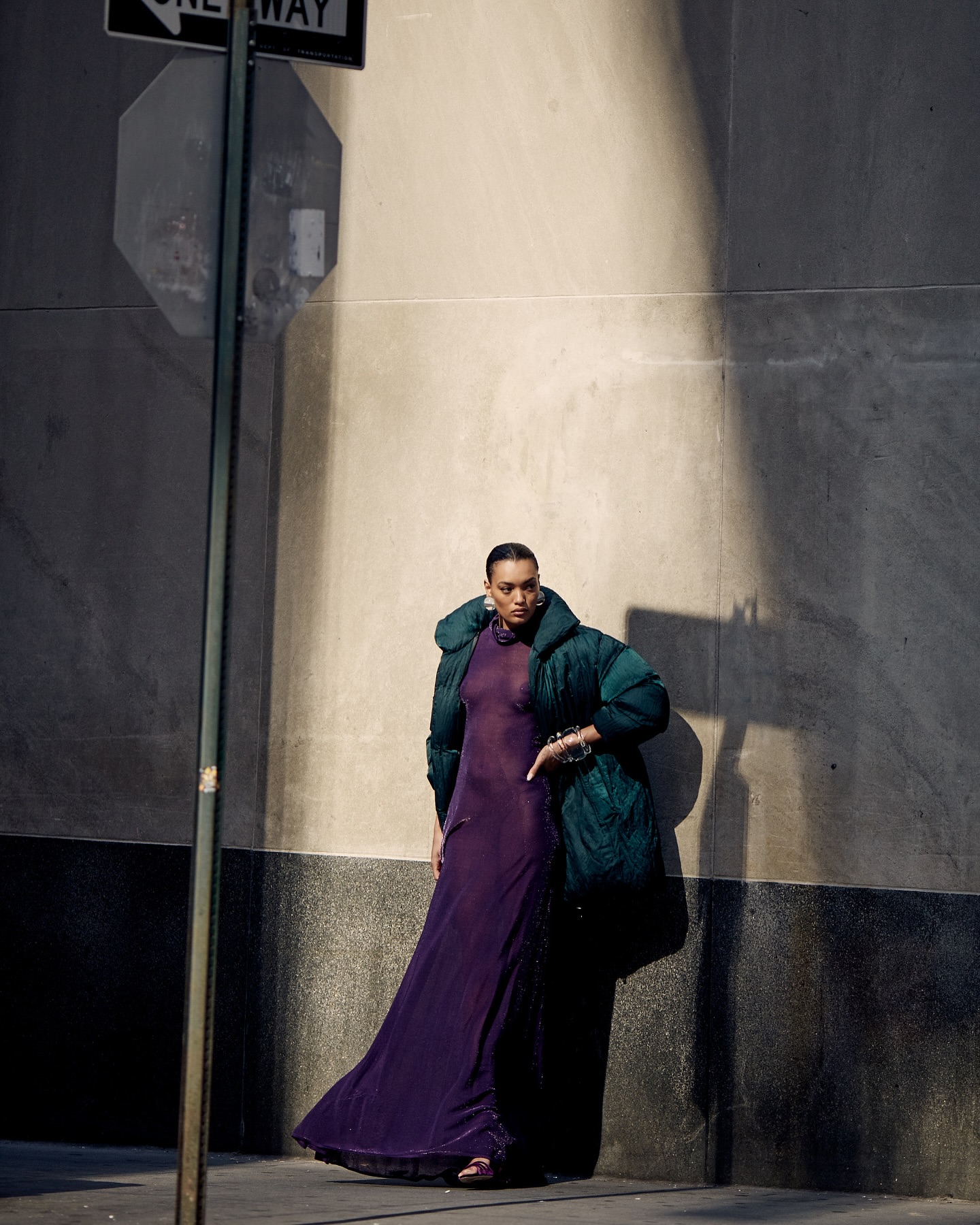 Fashionable woman in a purple gown and green puffer coat poses against a concrete wall on a city street with a one-way sign overhead.
