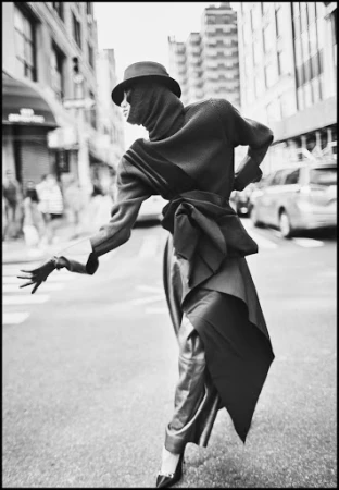 Model in a dark hat, hooded sweater, and draped wrap skirt walking along a city street in a monochrome urban editorial photograph.