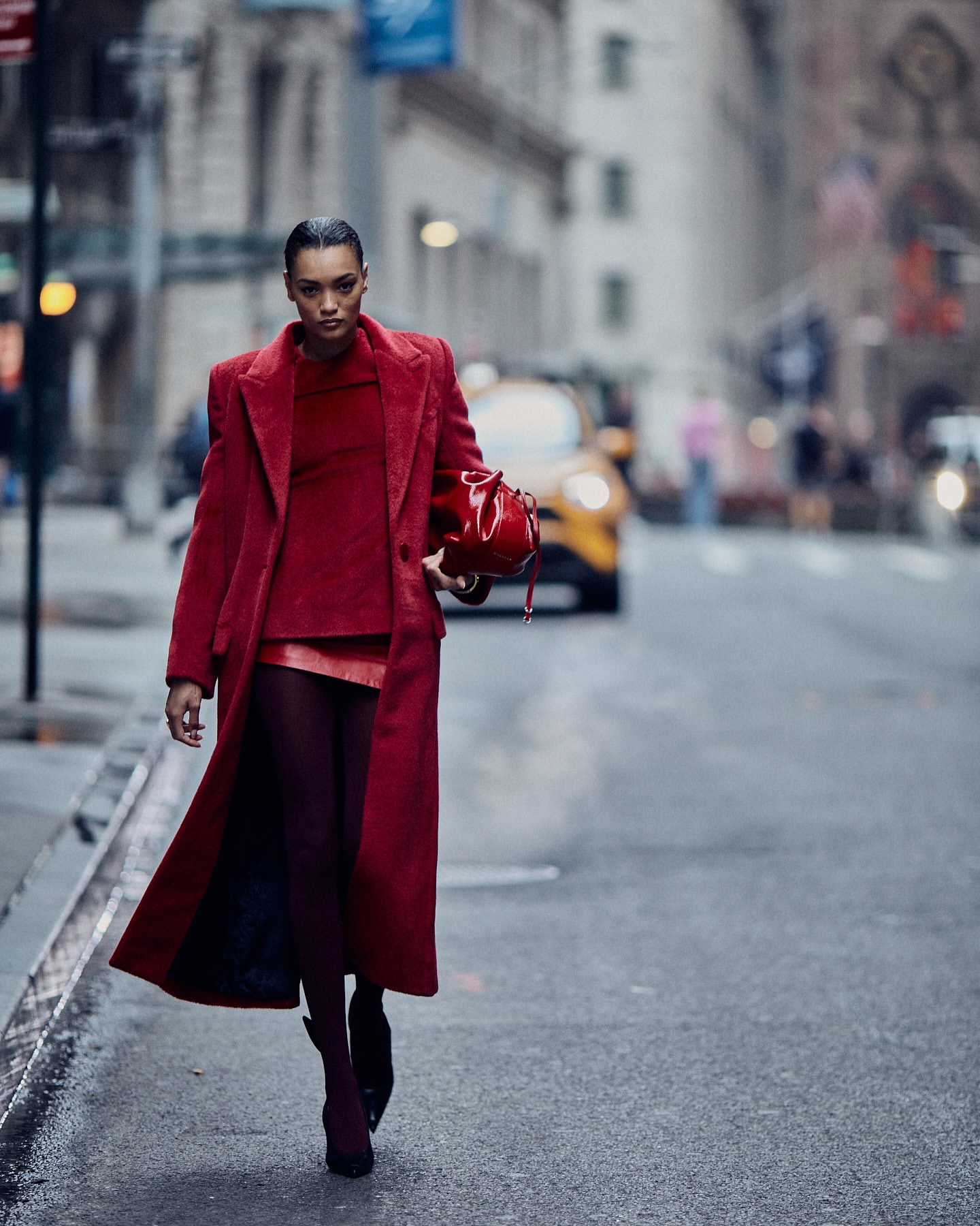 A woman in a red coat walks on a city street carrying a matching red purse. A yellow taxi is blurred in the background.