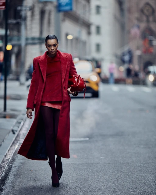 A woman in a red coat walks on a city street carrying a matching red purse. A yellow taxi is blurred in the background.