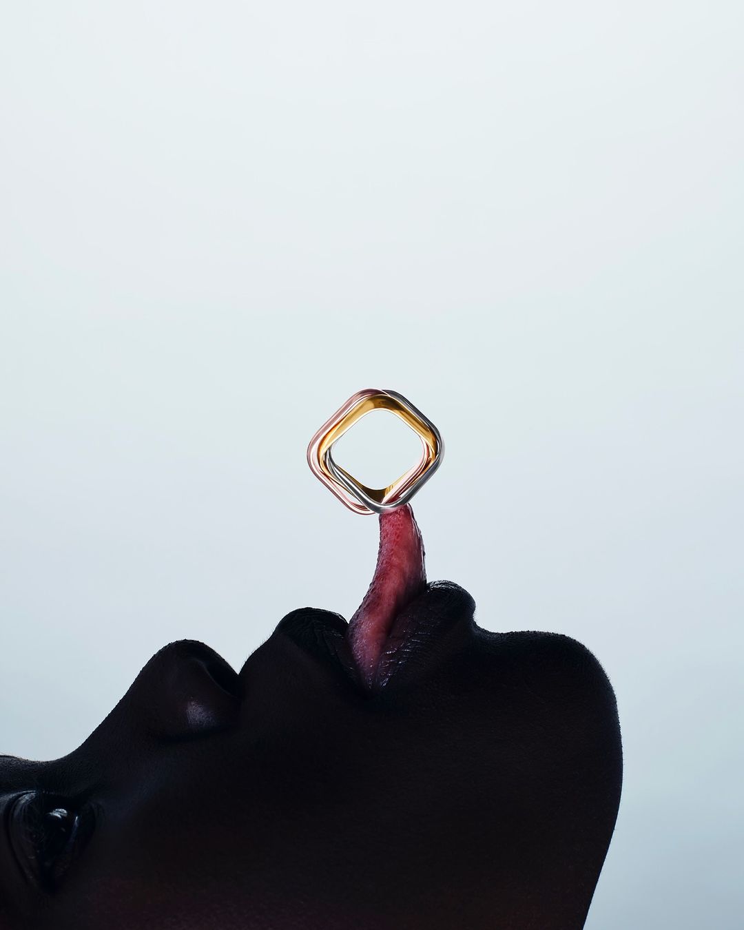 Close-up of a person's lips and tongue with a shiny, geometric ring balanced on the tongue against a pale background in a studio setting.