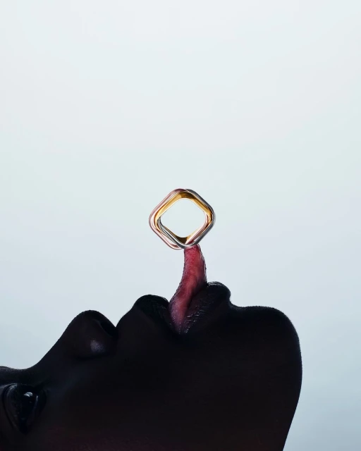Close-up of a person's lips and tongue with a shiny, geometric ring balanced on the tongue against a pale background in a studio setting.