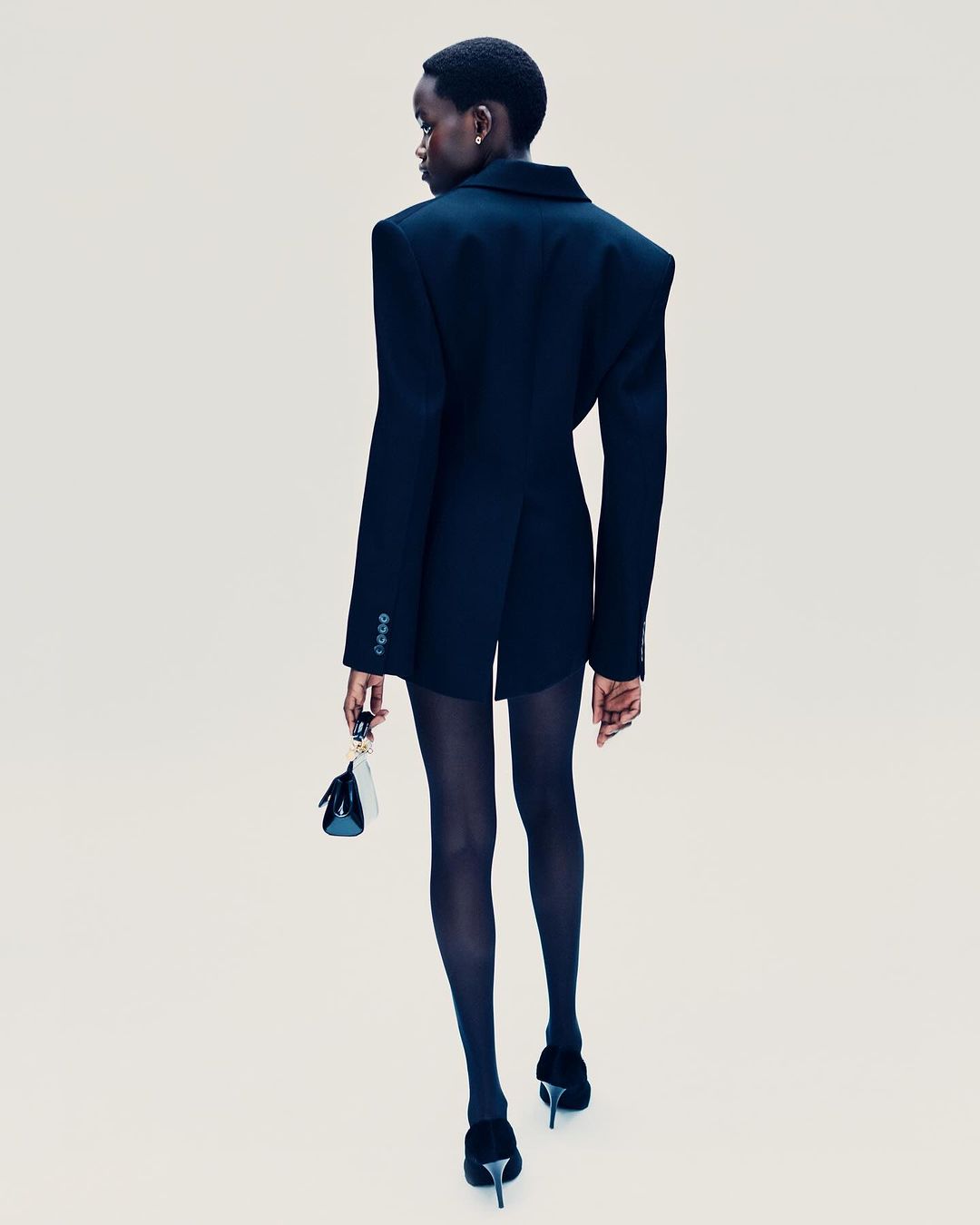 A slender, dark-skinned model in a black blazer dress, dark stockings, and heels, carries a mini handbag against a plain backdrop.