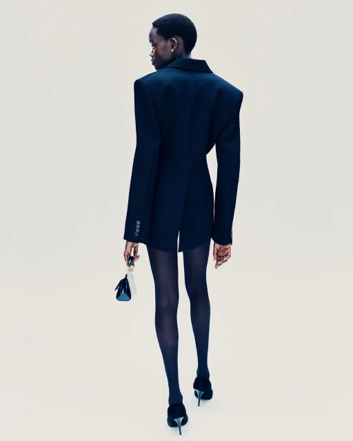 A slender, dark-skinned model in a black blazer dress, dark stockings, and heels, carries a mini handbag against a plain backdrop.
