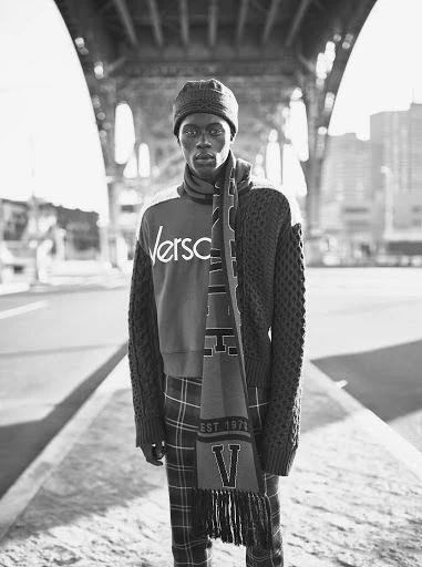 Black and white shot of an adult male wearing a Versace sweatshirt, scarf, and hat standing under a bridge in an urban setting.