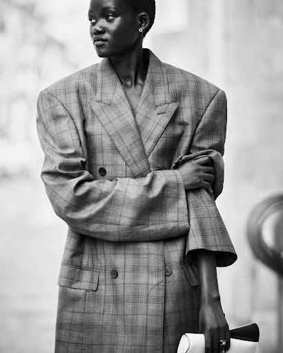 Black and white shot of a Black woman in an oversized plaid blazer holding a clutch. Minimalist jewelry. Blurred background.