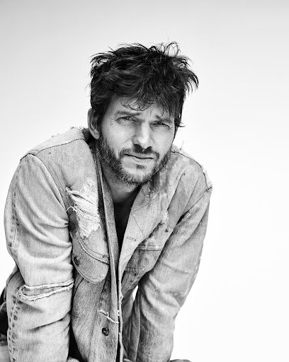 Black and white portrait of a man with tousled dark hair and a beard, wearing a distressed denim jacket, looking directly at the camera.
