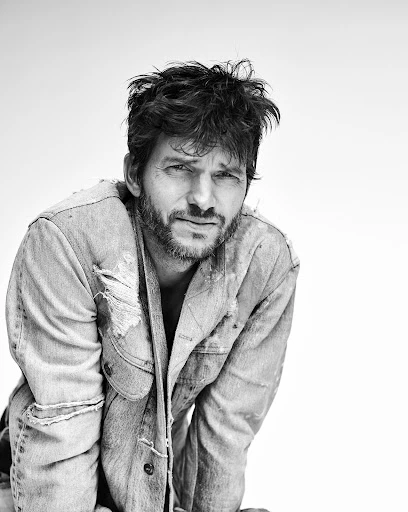 Black and white portrait of a man with tousled dark hair and a beard, wearing a distressed denim jacket, looking directly at the camera.