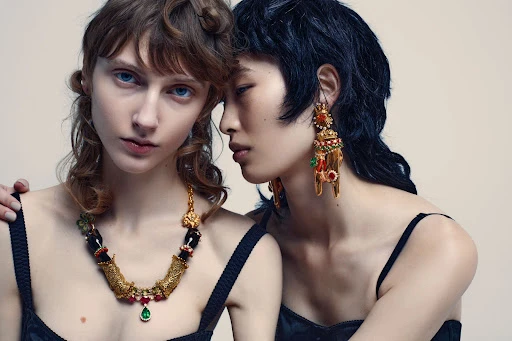 Two female fashion models pose in a studio setting with jewelry. One woman has auburn hair and a statement necklace while the other wears earrings.