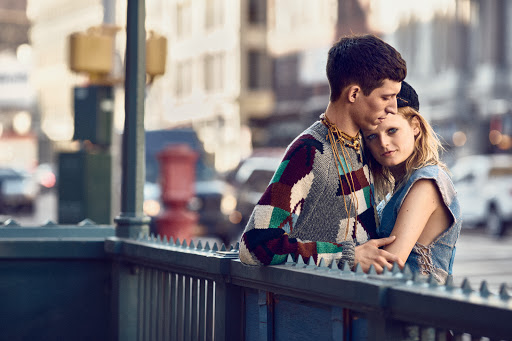 A stylish young couple poses near a subway entrance, the woman resting her head on the man's shoulder in an urban fashion shoot.
