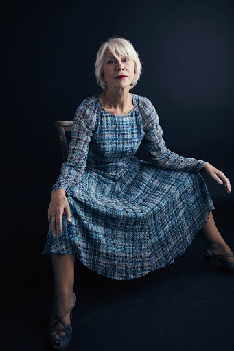 Helen Mirren seated on a dark wooden chair wearing a long-sleeved blue plaid midi dress against a dark background