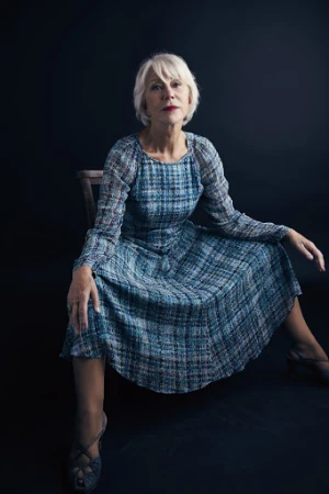 Helen Mirren seated on a dark wooden chair wearing a long-sleeved blue plaid midi dress against a dark background