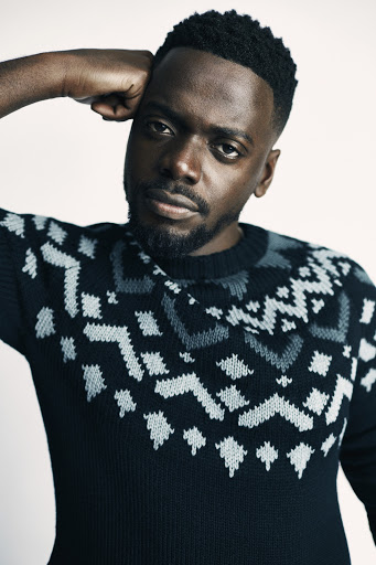 Portrait of an adult male wearing a black and grey patterned sweater indoors with a neutral background, posing and looking at the camera.