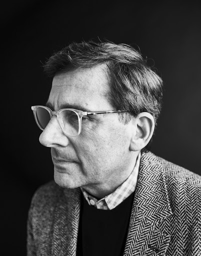 Portrait of a man wearing clear-rimmed glasses and a herringbone blazer against a dark background