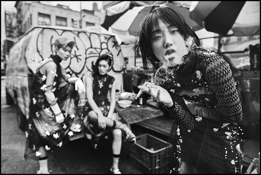 Three women in fashionable dresses pose in a black-and-white urban setting, with one smoking. The scene exudes a vintage and edgy vibe.
