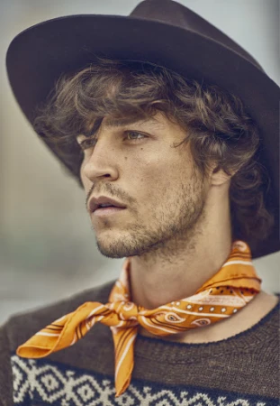Portrait of a man with curly brown hair wearing a dark fedora hat and an orange patterned bandana.