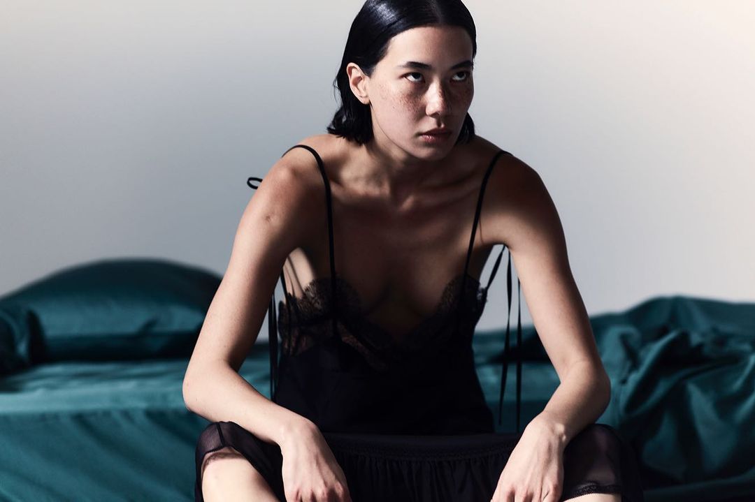 Elegant woman in a black slip dress seated on a bed in a photo studio with soft, diffused lighting. The colors are subdued and muted.