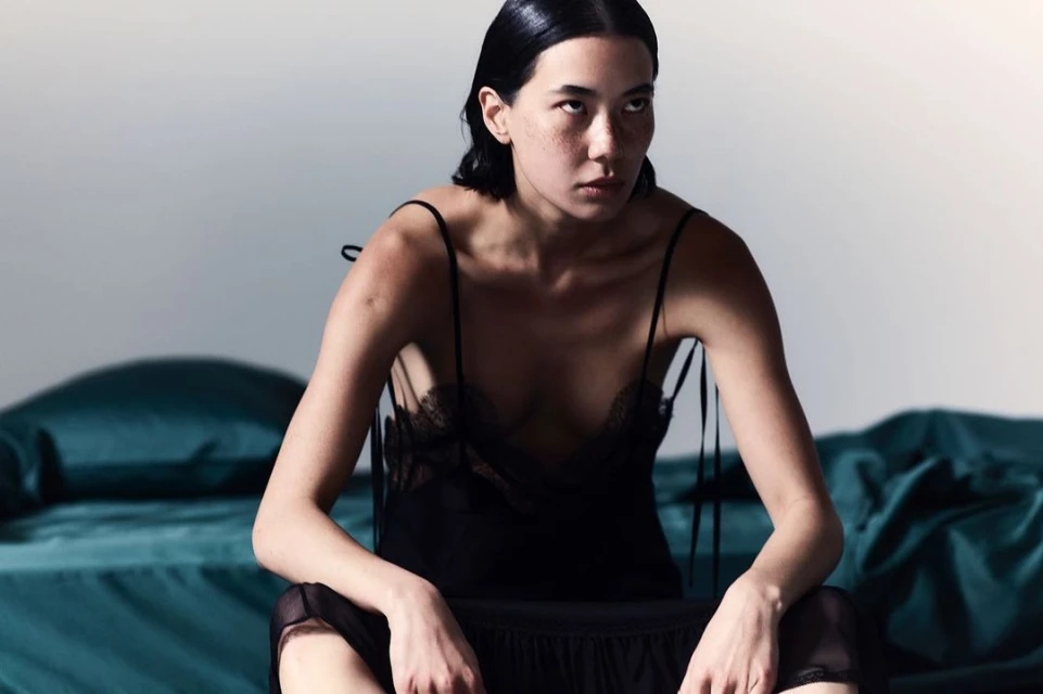 Elegant woman in a black slip dress seated on a bed in a photo studio with soft, diffused lighting. The colors are subdued and muted.
