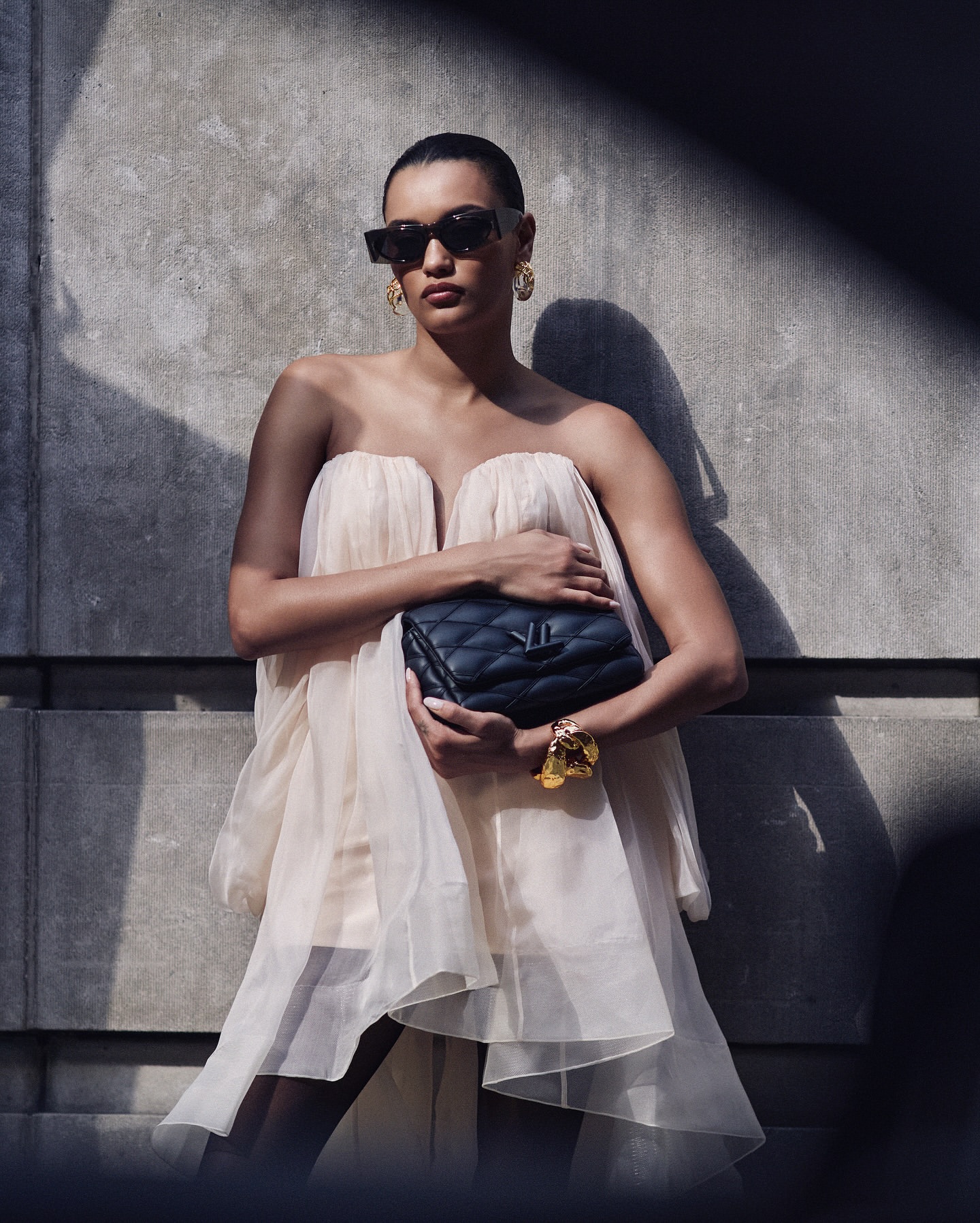 Stylish woman in a strapless dress and sunglasses holds a black handbag against a textured concrete wall with shadows, fashion shoot.