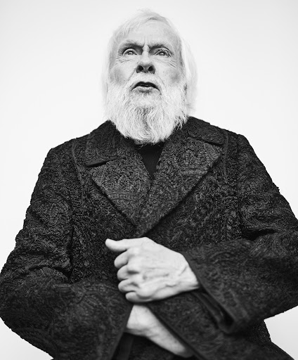 Black and white portrait of a senior male with a white beard and hair wearing a patterned black coat against a white backdrop.