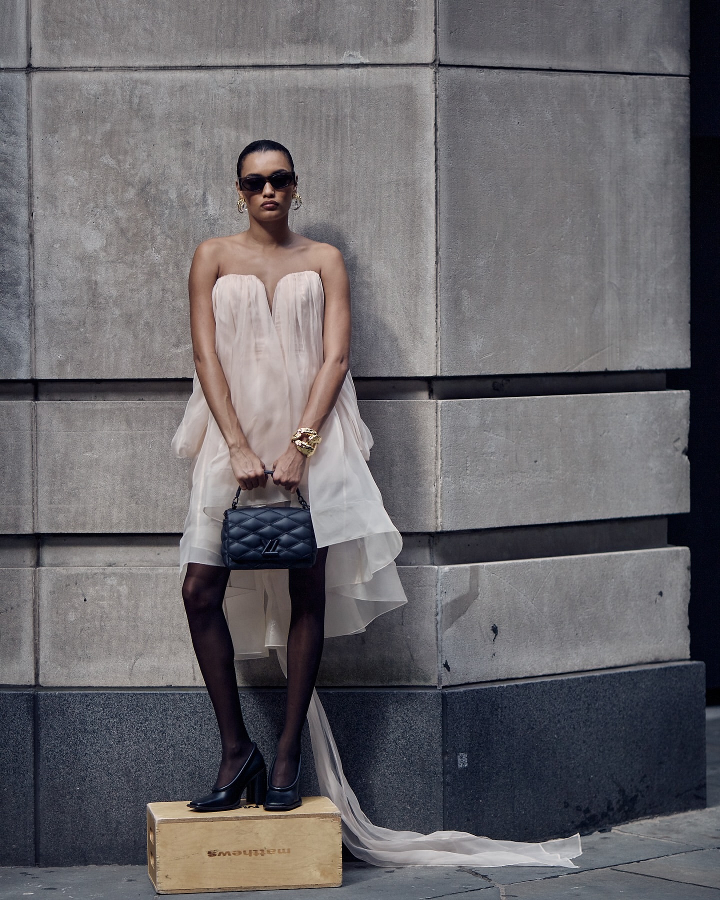 Woman in a strapless, flowing dress with a quilted handbag, gold bracelets, sunglasses and black heels posing against a concrete wall.