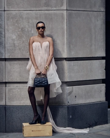 Woman in a strapless, flowing dress with a quilted handbag, gold bracelets, sunglasses and black heels posing against a concrete wall.
