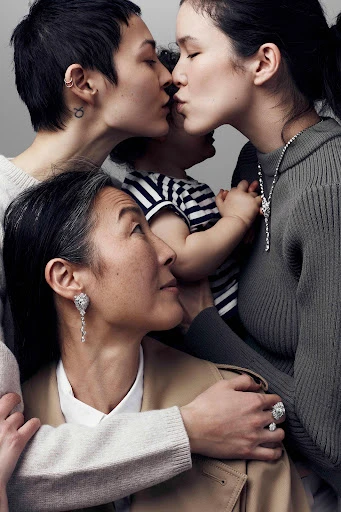 Three generations of women including an Asian grandmother, mother, and daughter embrace while showcasing fine jewelry in a neutral studio.