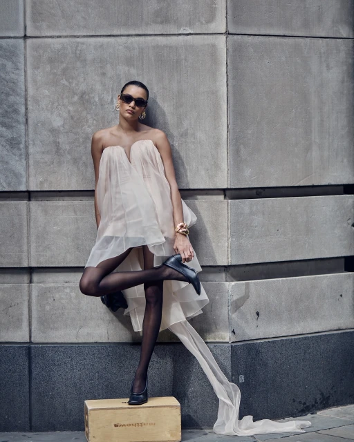 Woman in sheer peach dress and black tights poses against concrete wall, wearing jewelry and heels, with wooden box prop.