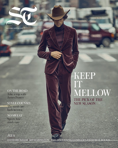 Magazine cover featuring a male fashion model in a brown velvet suit and hat, walking on a city street with traffic in the background.