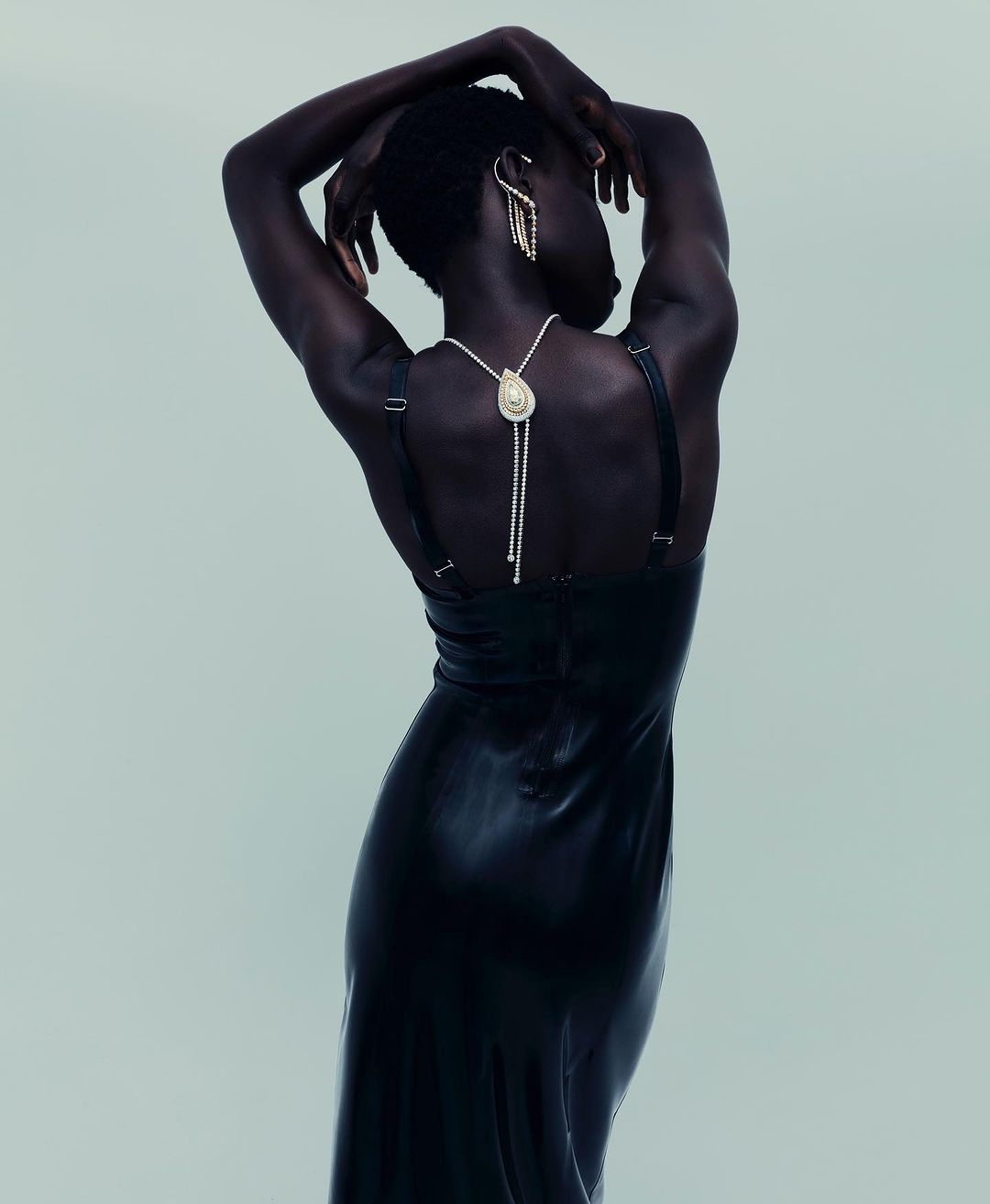 Elegant woman with dark skin poses in a long black dress with ornate jewelry against a light background, showcasing fashion and beauty.