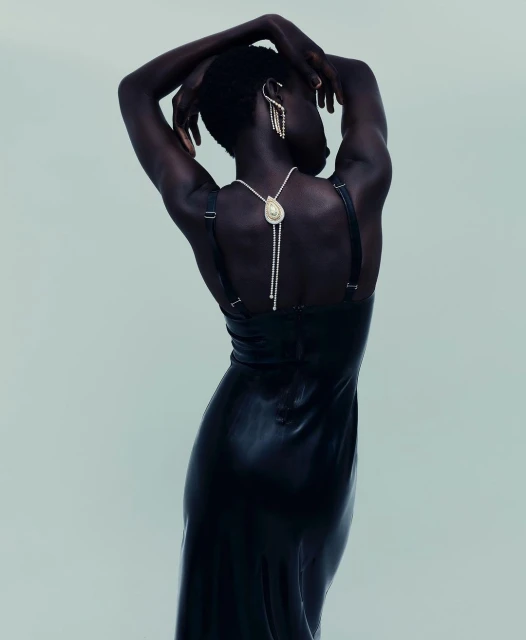 Elegant woman with dark skin poses in a long black dress with ornate jewelry against a light background, showcasing fashion and beauty.