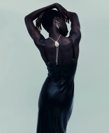 Elegant woman with dark skin poses in a long black dress with ornate jewelry against a light background, showcasing fashion and beauty.