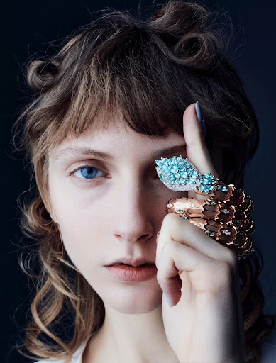 Close-up of a fair-skinned young adult woman with curly brown hair and blue eyes posing with snake-motif jewelry on her hand.