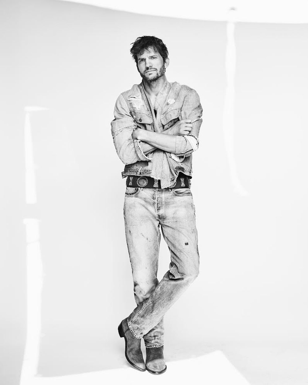 Man standing with arms crossed wearing a distressed denim shirt and jeans against a seamless white background.