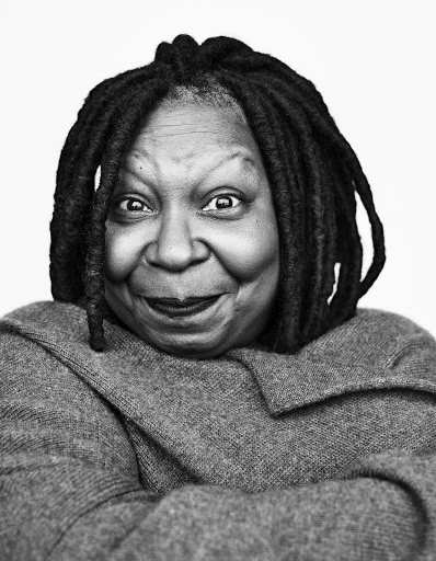 Monochrome portrait of Whoopi Goldberg, an adult female with dreadlocks, smiling in a studio setting, wearing a gray sweater.
