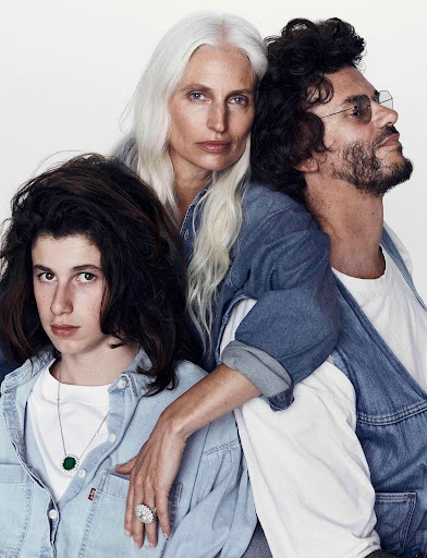 Three people pose indoors against a white background: a young adult, a mature woman with long grey hair, and a man with dark curly hair.