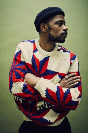 Portrait of a man with a beard wearing a patterned knit sweater and black beanie, photographed by Billy Kidd against a green backdrop.