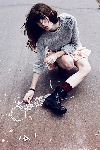 Young woman in a gray sweater and pink dress crouches on asphalt drawing chalk figures, accessorized with socks and black combat boots.