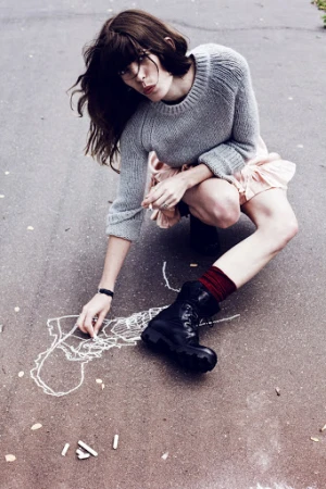 Young woman in a gray sweater and pink dress crouches on asphalt drawing chalk figures, accessorized with socks and black combat boots.