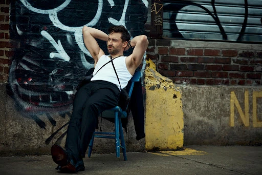An adult man poses on a blue chair in front of a graffiti-covered wall outside. He is wearing dress pants, a tank top, and suspenders.