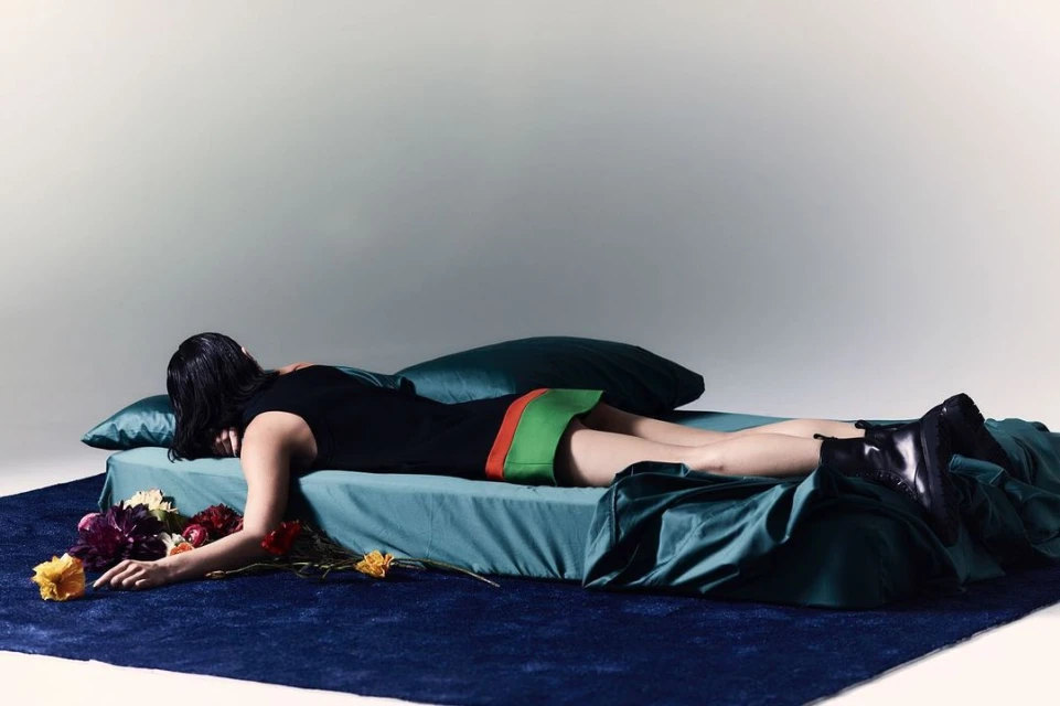 Adult woman lying on a teal-colored bed with flowers, wearing a black dress and boots against a light grey background in a studio shot.